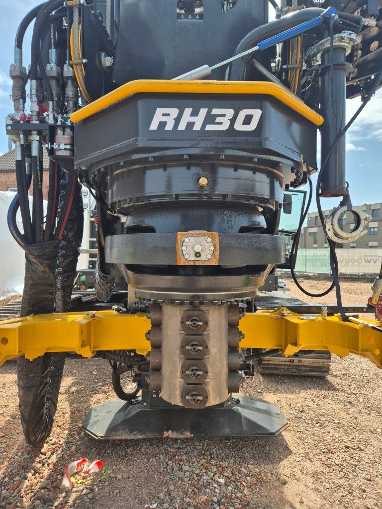 Close-up of a RH30 hydraulic breaker attachment mounted on a yellow support frame at a construction site, with hoses and metal components visible above.