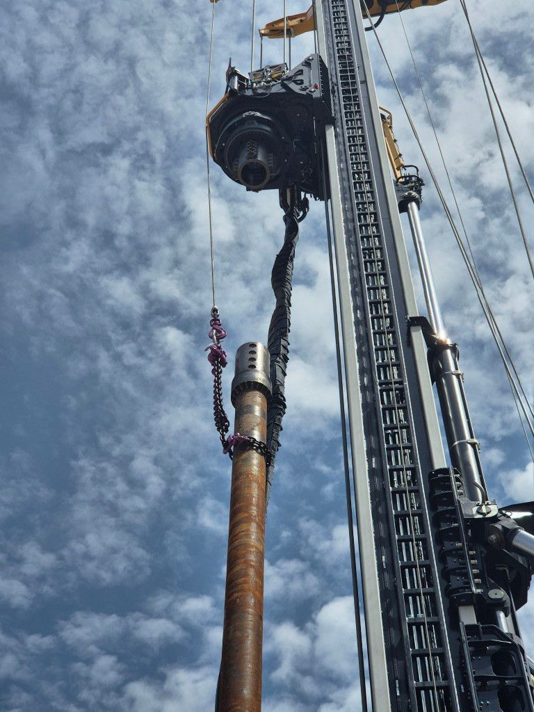 Drilling rig with a long steel drill pipe and lifting cables against a blue sky.