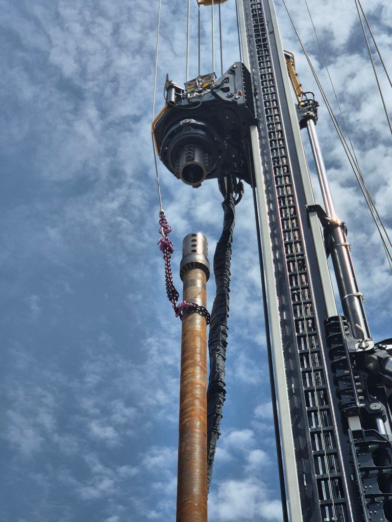 Heavy drilling equipment with a large brown drill pipe suspended by a crane against a blue sky; focus on the drill string and hoisting setup.