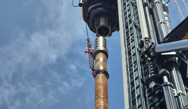 Industrial drilling rig lifting a long, rust-colored drill pipe into a borehole against a blue sky backdrop
