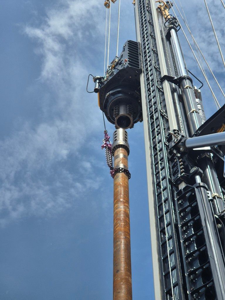 Industrial drilling rig lifting a long, rust-colored drill pipe into a borehole against a blue sky backdrop