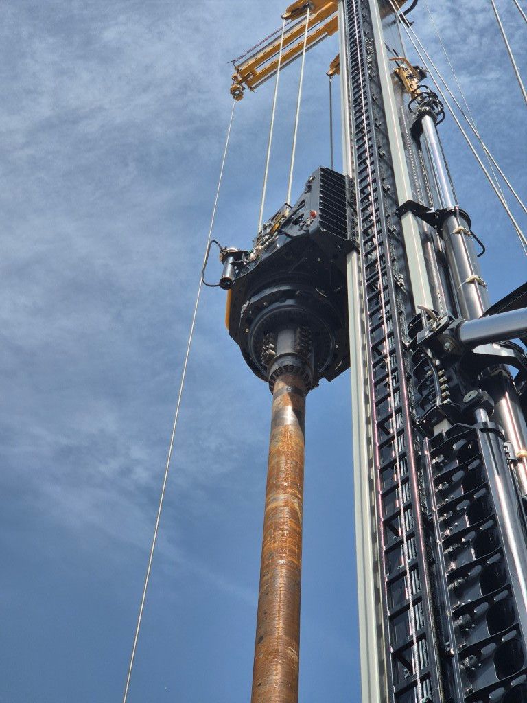 Drilling rig with a long steel drill pipe extending upward against a blue sky.